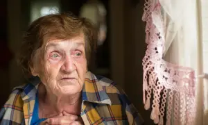Starsza kobieta zamyślona, z rękami na piersi, przy oknie z koronkową firanką. W jej oczach widać smutek, być może to depresja starcza.