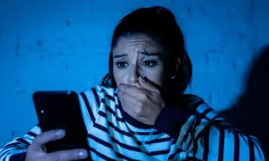 Przerażona kobieta trzyma telefon, jej dłoń zakrywa usta. Czyta wiadomość, która jest ewidentnym szantażem.