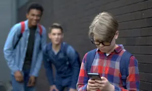 Chłopiec w okularach patrzy w telefon, podczas gdy dwaj inni chłopcy śmieją się za nim. Stop hejt!