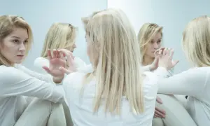Kobieta w białej bluzce patrzy na swoje odbicia w lustrze, które zdają się ją otaczać. Obraz sugeruje psychozę reaktywną i zagubienie.