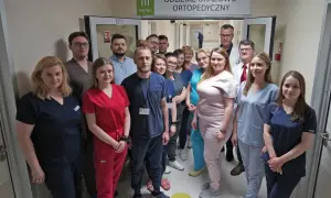 Grupa lekarzy i pielęgniarek w uniformach stoi w korytarzu szpitalnym pod znakiem "Oddział Urazowo-Ortopedyczny".