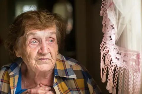 Starsza kobieta zamyślona, z rękami na piersi, przy oknie z koronkową firanką. W jej oczach widać smutek, być może to depresja starcza.