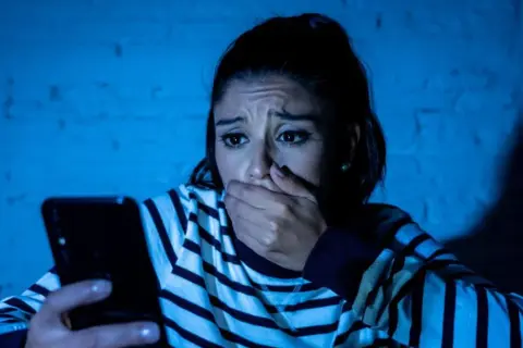 Przerażona kobieta trzyma telefon, jej dłoń zakrywa usta. Czyta wiadomość, która jest ewidentnym szantażem.
