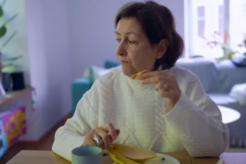 Kobieta w białym swetrze je banana, czując uczucie guli w gardle.
