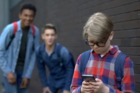 Chłopiec w okularach patrzy w telefon, podczas gdy dwaj inni chłopcy śmieją się za nim. Stop hejt!