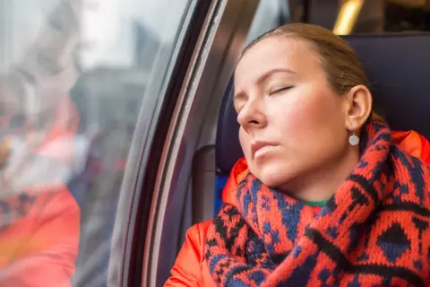 Kobieta w pomarańczowej kurtce i wzorzystym szaliku odpoczywa w pociągu, wyraźnie odczuwając zmęczenie.