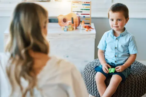 Chłopiec w niebieskiej koszuli siedzi na pufie, rozmawiając z psychoterapeutką.