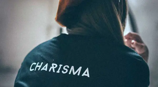 Osoba w bluzie z napisem "CHARISMA" na plecach, z włosami opadającymi na ramiona i dłonią przy uchu.