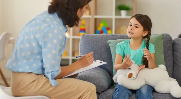 Psychoterapeutka rozmawia z dziewczynką trzymającą misia w gabinecie.