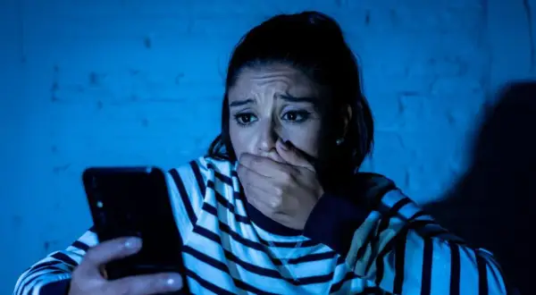 Przerażona kobieta trzyma telefon, jej dłoń zakrywa usta. Czyta wiadomość, która jest ewidentnym szantażem.