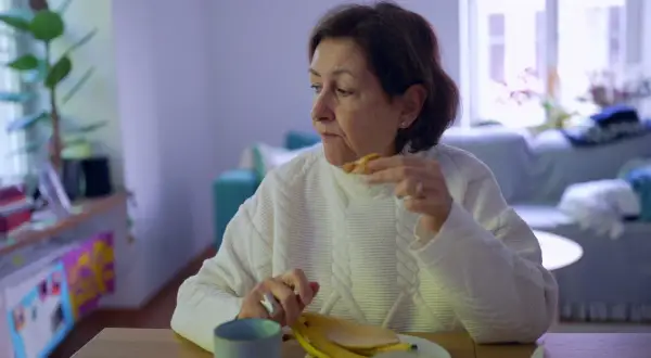 Kobieta w białym swetrze je banana, czując uczucie guli w gardle.