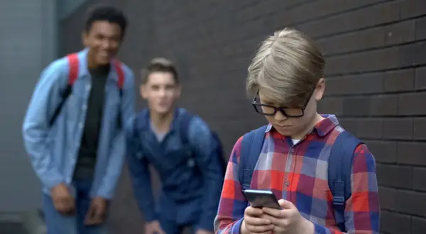 Chłopiec w okularach patrzy w telefon, podczas gdy dwaj inni chłopcy śmieją się za nim. Stop hejt!