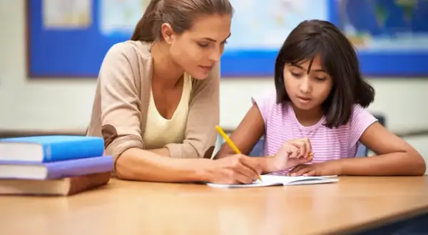 Nauczycielka pomaga uczennicy w pisaniu, wspierając ją w procesie edukacyjnym, który może być częścią orzeczenia z poradni psychologiczno-pedagogicznej.