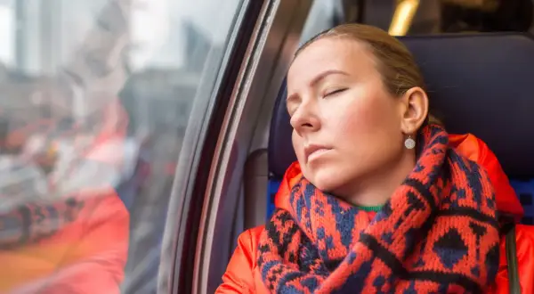 Kobieta w pomarańczowej kurtce i wzorzystym szaliku odpoczywa w pociągu, wyraźnie odczuwając zmęczenie.