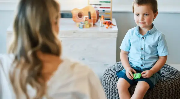 Chłopiec w niebieskiej koszuli siedzi na pufie, rozmawiając z psychoterapeutką.