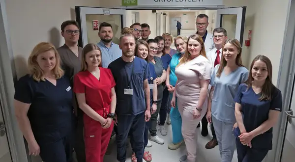 Grupa lekarzy i pielęgniarek w uniformach stoi w korytarzu szpitalnym pod znakiem "Oddział Urazowo-Ortopedyczny".