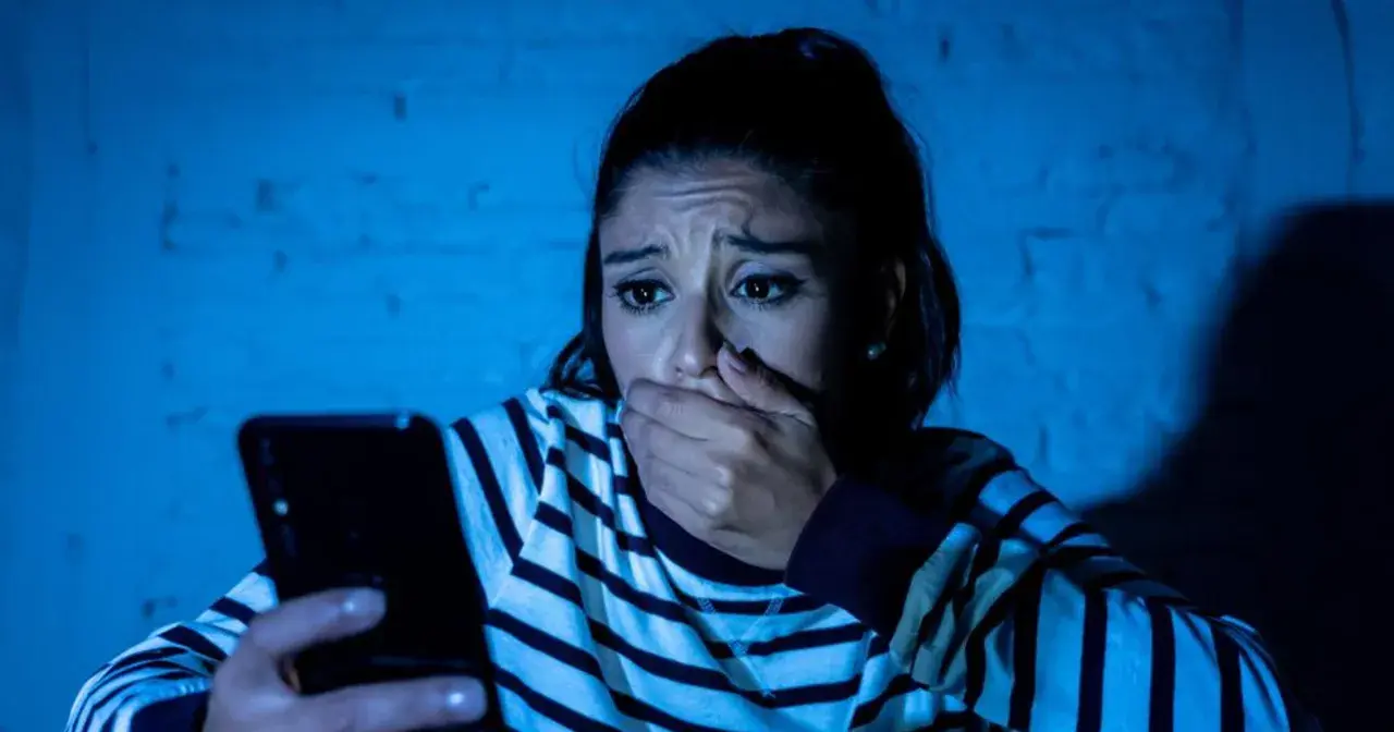 Przerażona kobieta trzyma telefon, jej dłoń zakrywa usta. Czyta wiadomość, która jest ewidentnym szantażem.