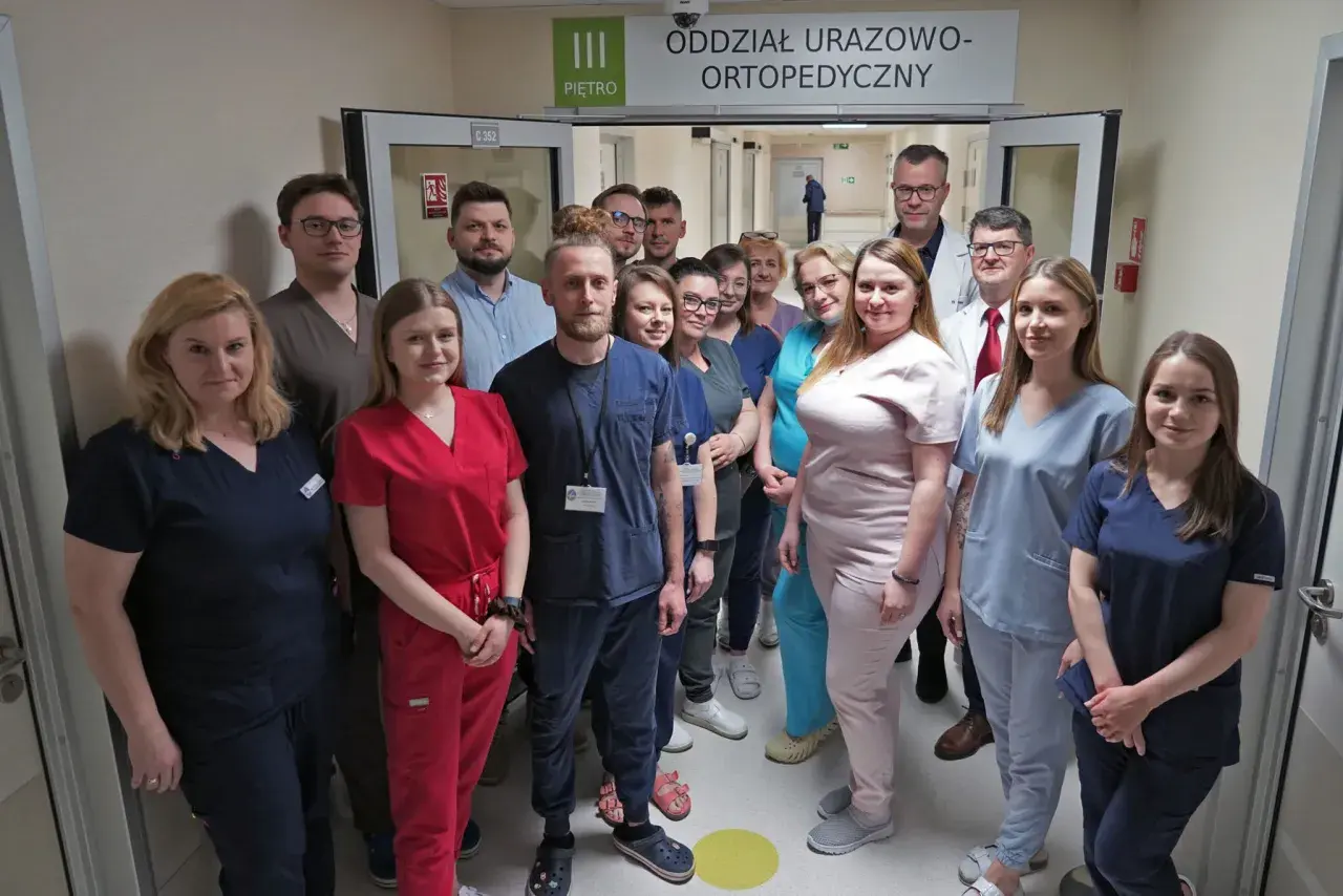 Grupa lekarzy i pielęgniarek w uniformach stoi w korytarzu szpitalnym pod znakiem "Oddział Urazowo-Ortopedyczny".