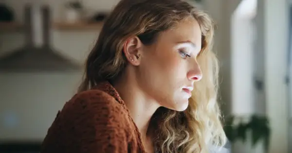 Młoda kobieta z blond włosami, w brązowym swetrze, patrzy w dal. Jej wyraz twarzy sugeruje dysocjację, jakby była nieobecna myślami.