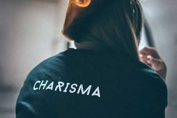 Osoba w bluzie z napisem "CHARISMA" na plecach, z włosami opadającymi na ramiona i dłonią przy uchu.