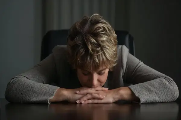Kobieta z opuszczoną głową, ręce splecione na stole. Wygląda na wyczerpaną, jakby przechodziła trudny test zmęczenia psychicznego.