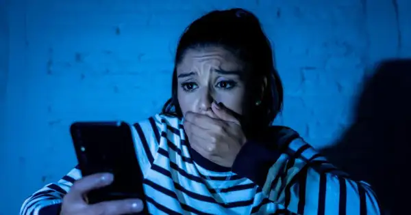 Przerażona kobieta trzyma telefon, jej dłoń zakrywa usta. Czyta wiadomość, która jest ewidentnym szantażem.