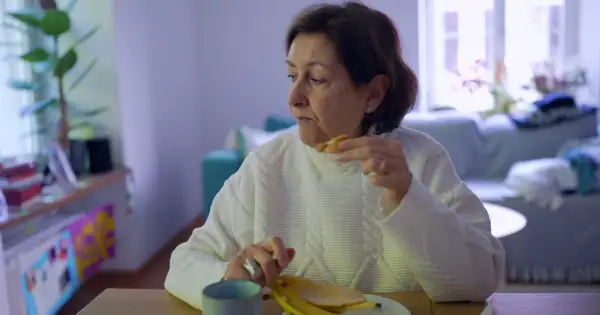 Kobieta w białym swetrze je banana, czując uczucie guli w gardle.