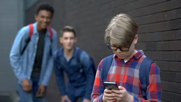 Chłopiec w okularach patrzy w telefon, podczas gdy dwaj inni chłopcy śmieją się za nim. Stop hejt!