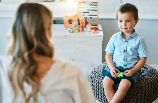 Chłopiec w niebieskiej koszuli siedzi na pufie, rozmawiając z psychoterapeutką.